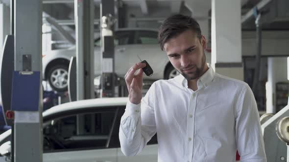 Young Handsome Man in Car Service Shop Showing His Car Keys and Smiling on the Background of His Car