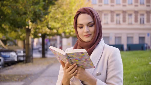 A Young Beautiful Muslim Woman Reads a Book with a Smile in a Street in an Urban Area  Closeup