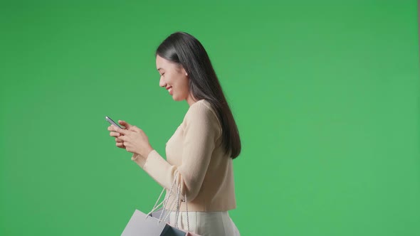 Side View Of Asian Woman With Shopping Bags Using Mobile Phone While Walking At Green Screen