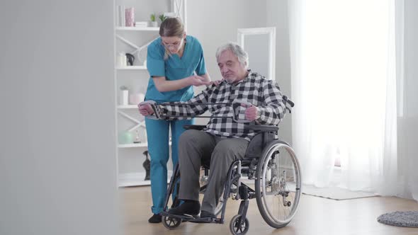 Long Shot of Concentrated Caucasian Woman Helping Old Man To Stretch Elastic Band. Mature Retiree