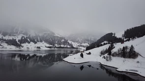 Flight around a small settlement of houses on a snowy hill next to a mountain lake in the winter in