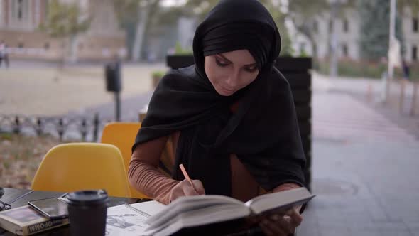 An Attractive Muslim Girl Writes in a Notebook Sitting in a Cafe with a Book