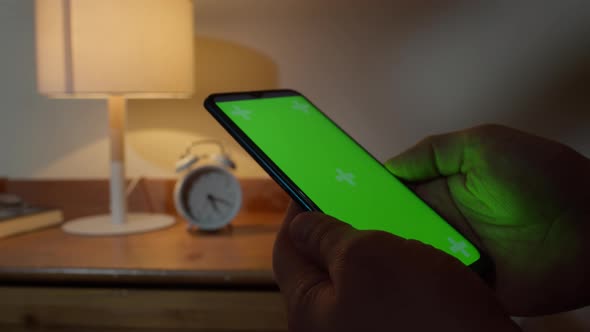 Man Sitting on a Sofa Using a Smartphone with a Colored Green Screen at Night