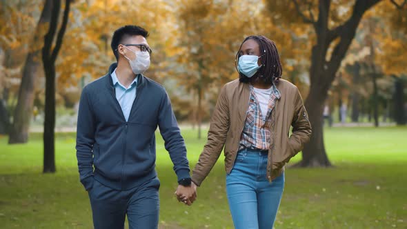 Multiethnic Couple in Protective Masks Holding Hands Walking in Park