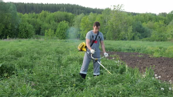 Man in Gray Work Clothes Mows Nettles with a Trimmer in the Field