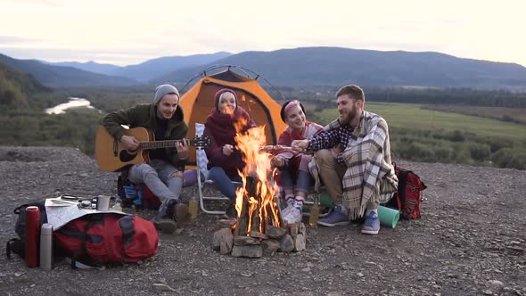 Young guy traveler is playing the guitar, his friends are cooking sausages on fire