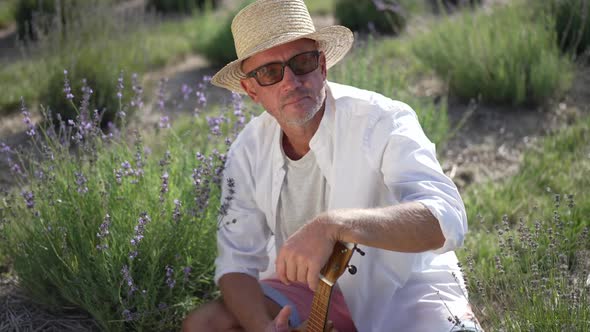 Portrait of Thoughtful Handsome Man with Ukulele Sitting on Lavender Field Looking Away