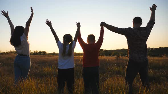 Mommy and Daddy with Two Small Children Stand on Grass Field with Raising Hands at Sunset