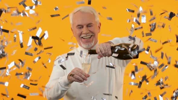 Smiling Senior Man Celebrating Holiday Pouring Champagne Under Falling Confetti