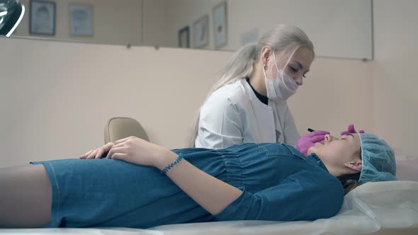 Brow Master in Gloves Works with Young Woman Lying on Couch