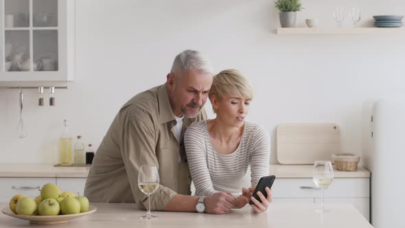 Discontented MiddleAged Couple Using Smartphone Scrolling Negative News In Kitchen