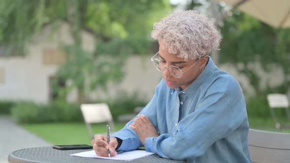 Young African Woman Writing on Paper in Outdoor Cafe