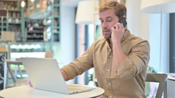 Young Man with Laptop Talking on Smartphone in Cafe