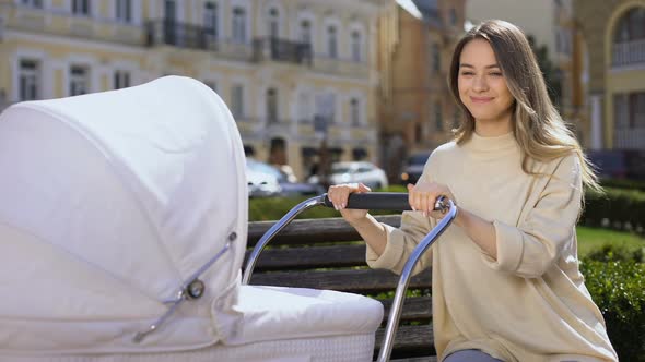 Young Happy Mother Admiring Infant Swinging Stroller Sitting on Park Bench