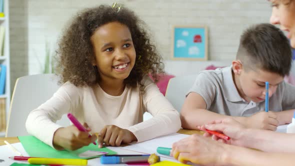 Little African Girl Drawing at Table in Kindergarten and Talking with Teacher