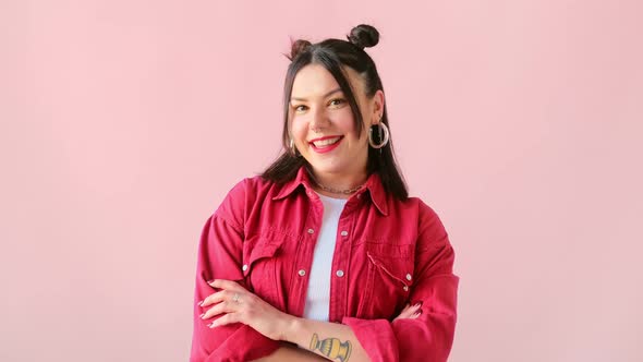 Stylish brunette woman with tattoo blinking eye, smiling and looking at camera on pink background.