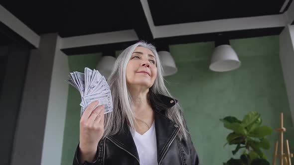 Satisfied Stylish Senior Woman which Waving Dollars Banknotes on the Room Background