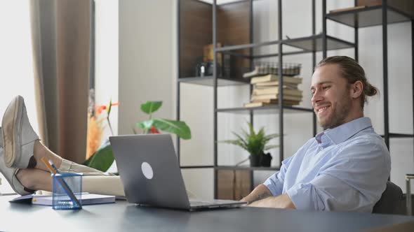 Smiling Attractive Young Man Freelancer or Manager is Relaxing at Workplace with Legs Thrown on