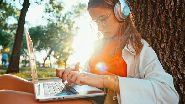 Fulllength Side View of Teenage Girl Sitting with Headphones in Park on Grass By Tree and Using