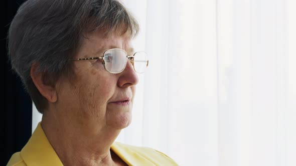 Closeup of the Face of an Old Woman with Glasses Who Stands Alone By the Window