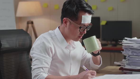 Close Up Of Sick Asian Man Drinking Water While Working Hard With Documents At The Office