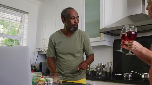 Diverse senior couple preparing food in kitchen using laptop and drinking wine