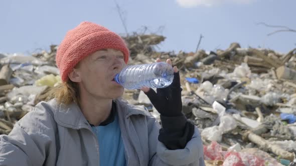 Homeless woman drinking water from plastic bottle in landfill