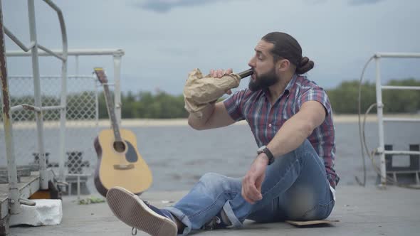 Sad Alcoholic Musician Sitting on Urban Embankment and Drinking Wine From Bottle in Paper Bag. Wide