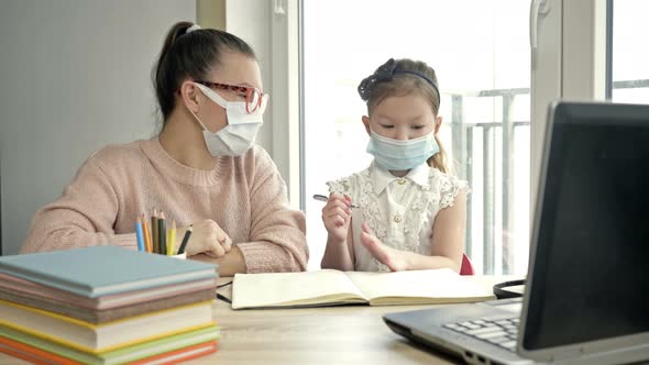 Mom or Teacher or Tutor Helps an Elementary School Student with Her Lessons. Both Have Medical Masks