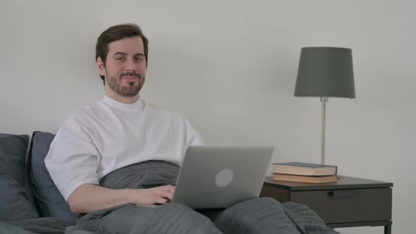 Young Man with Laptop Smiling at the Camera in Bed