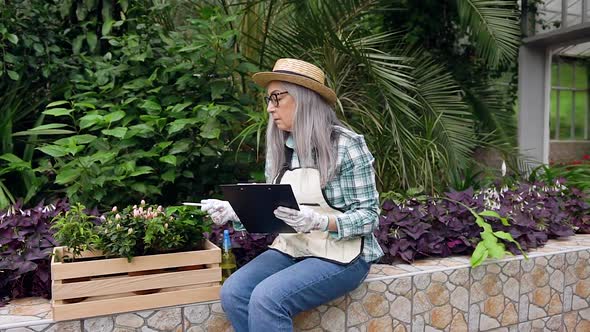 Female Gardener in Hat Sitting on Concrete Parapet and Working with Flowers and Documents