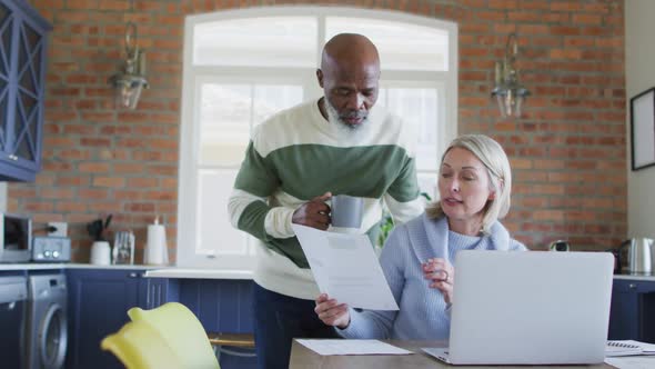 Stressed senior diverse couple in kitchen sitting at table, using laptop