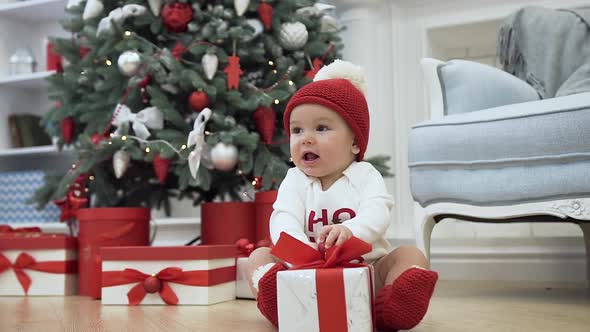 Cute Baby Dressed in the Christmas Costume and Red White Hat Sitting on the Wooden Floor