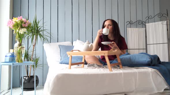 Young Attractive Woman in Pajamas Resting on Bed in Morning. Pretty Female Drinking Coffee Sitting