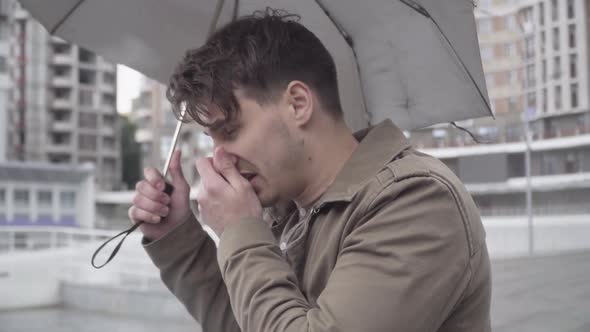 Close-up of Young Ill Caucasian Man Standing Outdoors with Umbrella, Sneezing and Coughing. Side