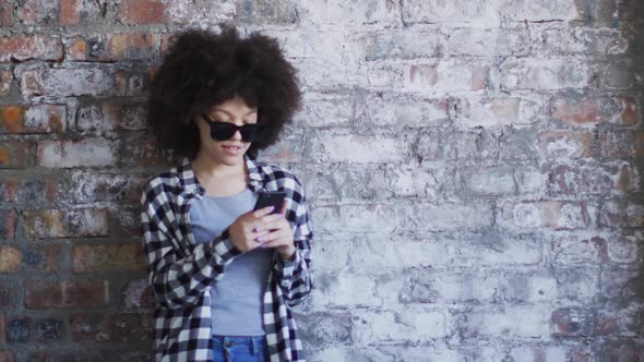 African american woman wearing sunglasses using smartphone while standing against brick wall