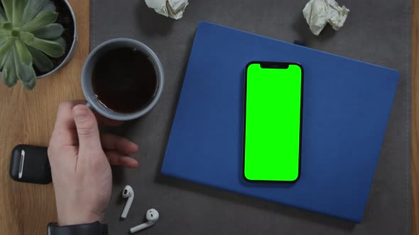 A Man Takes Cup of Coffee Standing on Desktop Next to Mobile with Green Screen