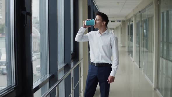 Young Man Drinks a Cup of Coffee and Looks Around Out the Windows in Corridor
