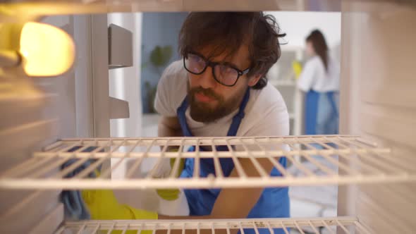 View From Inside Empty Refrigerator of Man Wearing Rubber Gloves and Apron Cleaning Shelves