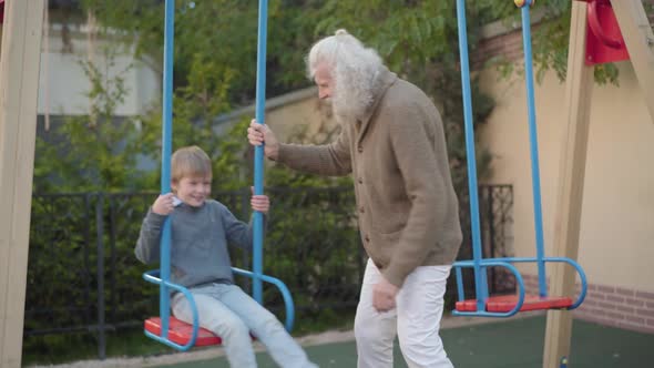 Senior Man Pushing Swings with Cute Smiling Boy Sitting in It. Portrait of Cheerful Caucasian