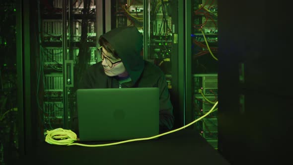 Asian male hacker in hoodie using laptop by computer servers