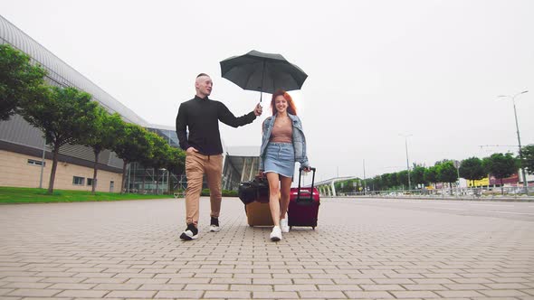 A Woman Carrying Two Suitcases To the Airport Terminal and a Man Holding an Umbrella. The Priority