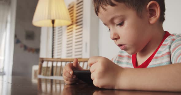 Portrait of a Little Boy Child with a Beautiful Face Looking at the Phone While Sitting at the Table