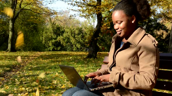 Young African Happy Girl Sits on Bench in Woods and Works on Laptop