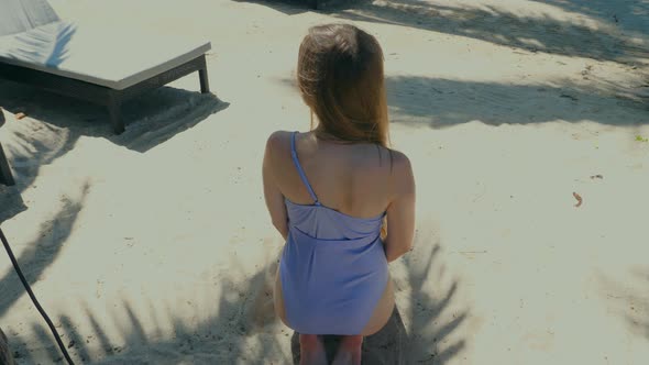 Back View of Beautiful Young Slim Woman in Swimsuit Sitting on Knees on White Sand Beach with