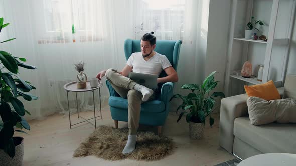 Young Man Using Laptop Typing and Working Sitting on the Armchair at Home and Drink Coffee