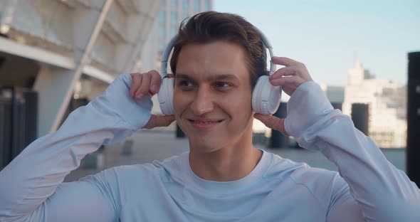 Portrait of a Sporty Man Runner Puts on Headphones Enjoys Music Before a Workout Standing Outdoors