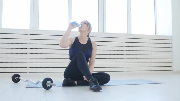 Fitness Concept. Young Woman Drinking Water After Sports Activities in the White Interior