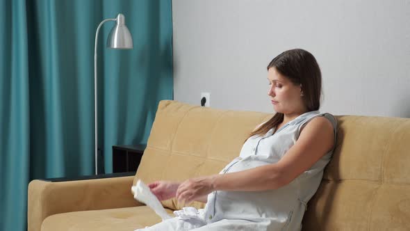 Pregnant Woman with Big Belly Prepares Baby Clothes While Sitting on Sofa