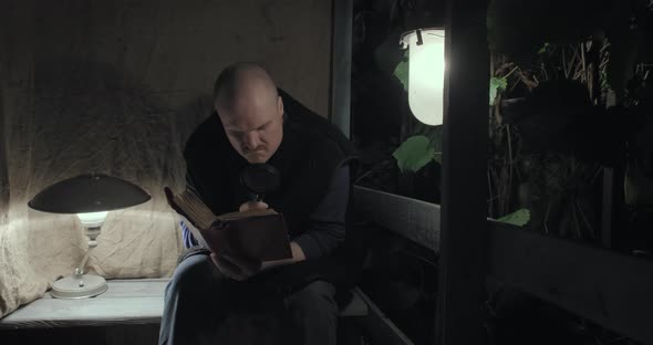 Elderly Angry Man Sits on Porch at Night and Reads a Book Using Magnifier Glass